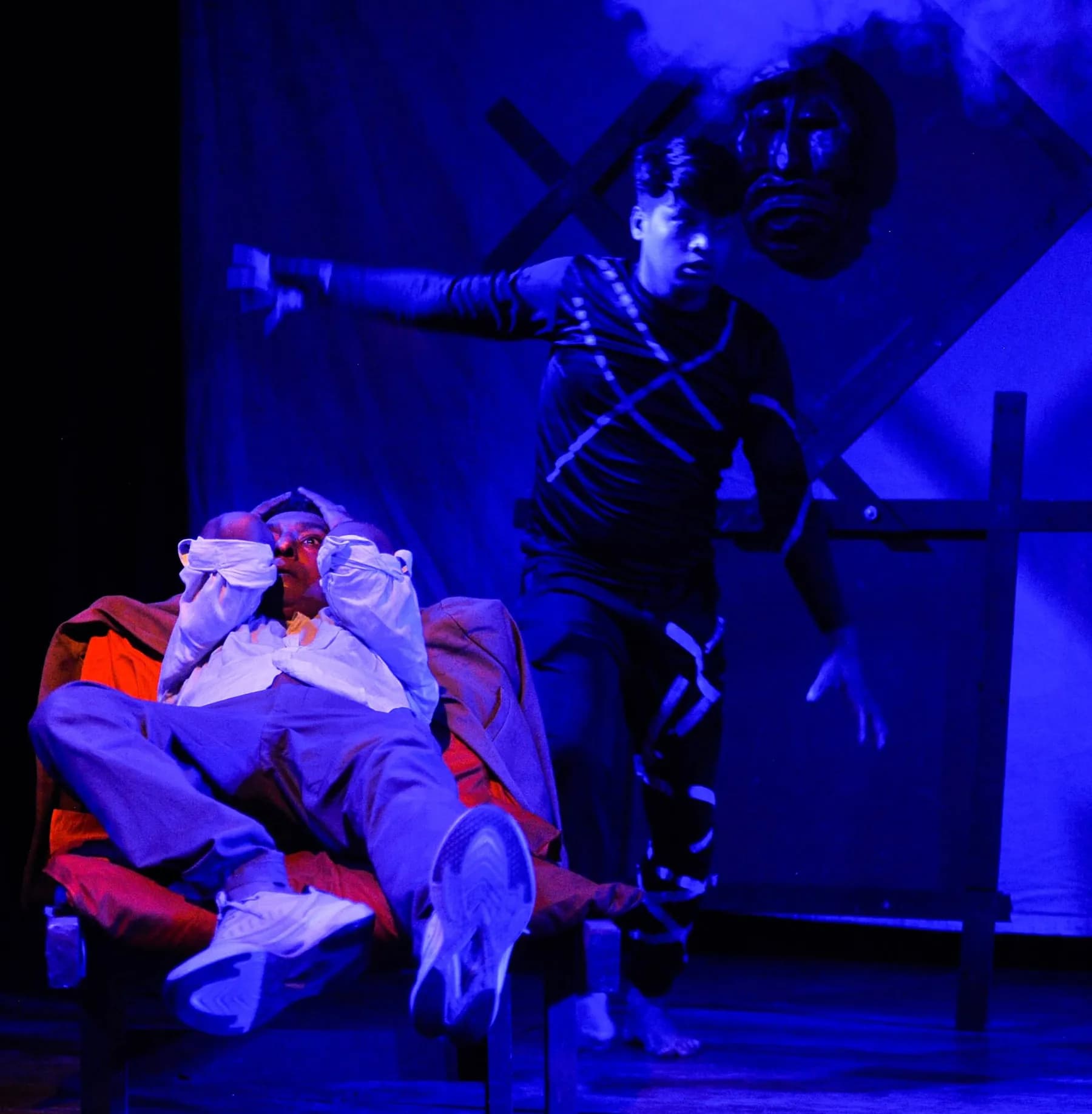 An actor in blue light beside a bound figure slumped in a chair