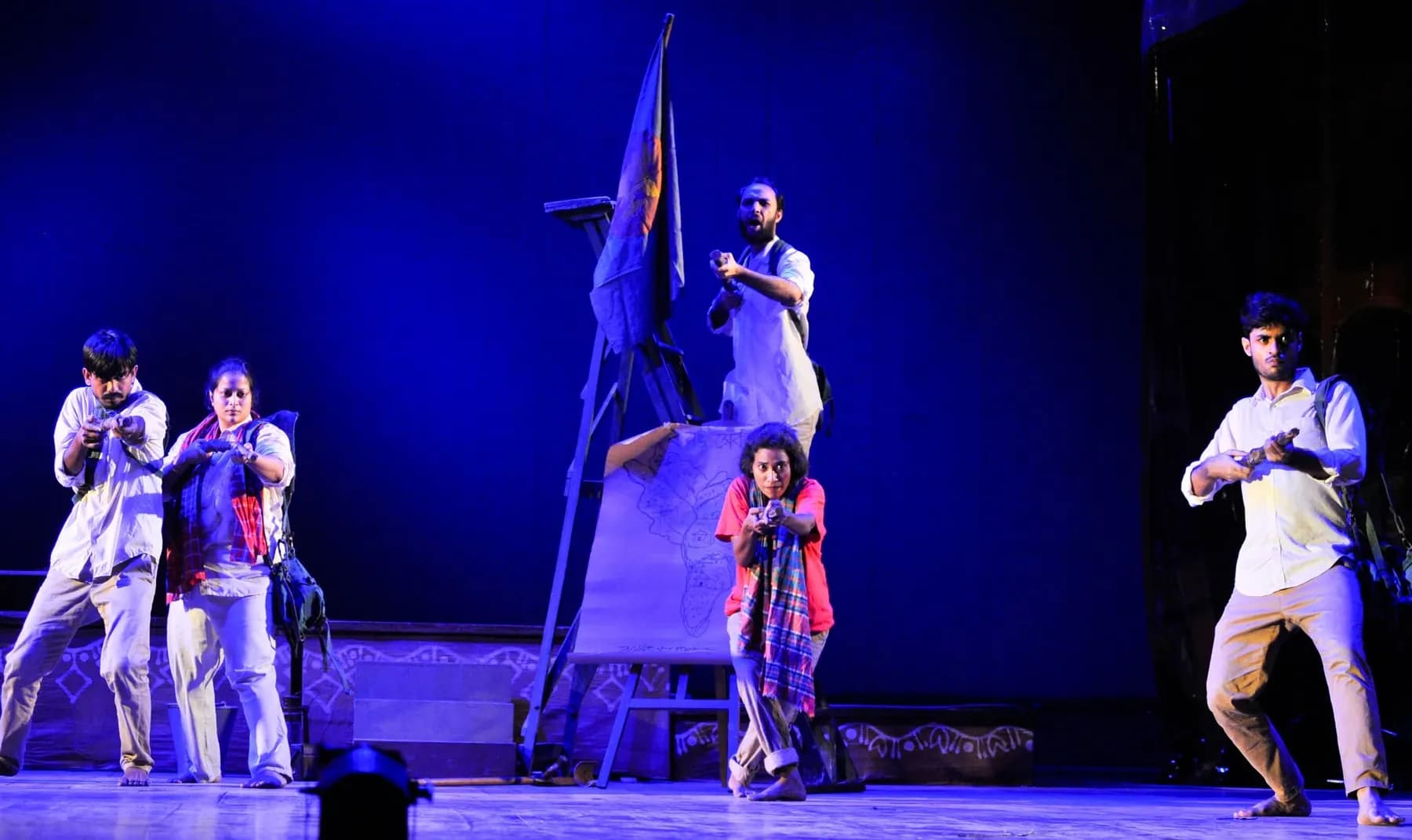 A company on stage under blue light, a flag raised behind them